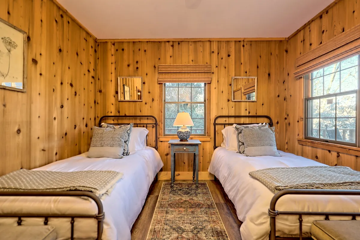 A twin bedroom with two inviting beds, wooden wall paneling, a table lamp, decorative pillows, and a window offering natural light. The setup feels cozy and perfect for relaxation or quiet conversation.