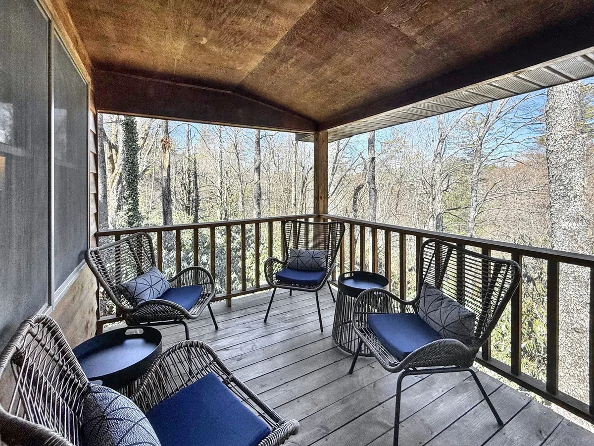 The image showcases the inviting exterior of a cozy cabin surrounded by lush natural greenery. A covered wooden deck features comfortable wicker chairs, perfect for relaxing and soaking in the tranquil mountain atmosphere. The deck's positioning offers an ideal spot for watching sunsets, birdwatching, and stargazing, all just steps from downtown Highlands.