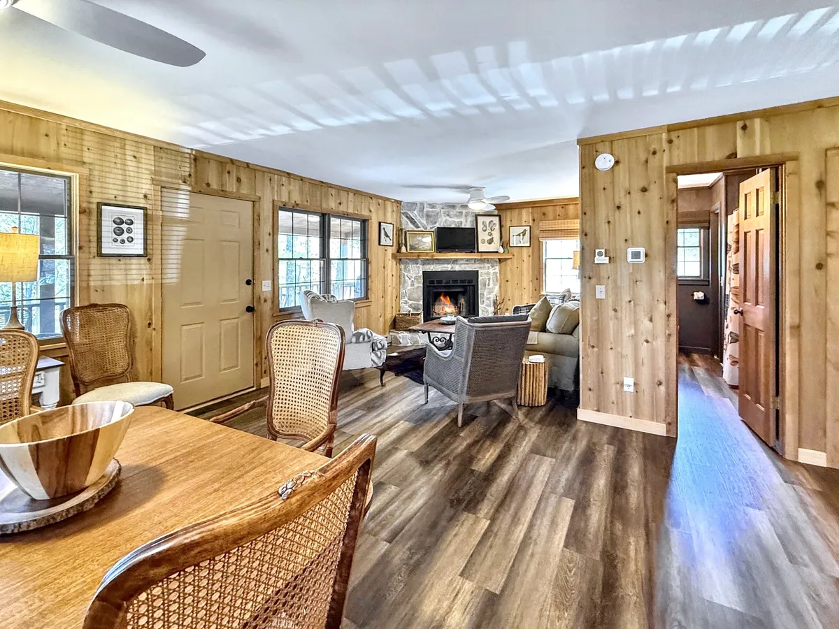 A welcoming open-concept interior showcasing a fireplace, a large dining table, and a cozy seating area. The room features wood-paneled walls and warm lighting, evoking a sense of comfort and togetherness—the perfect setting for family gatherings or relaxed evenings by the fire.