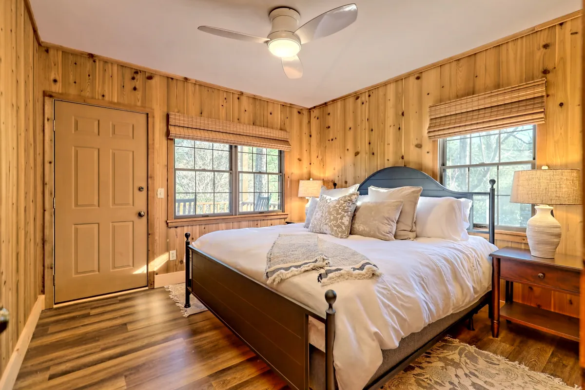 A serene bedroom with a cozy bed, windows letting in soft natural light, wooden walls, a nightstand, and warm light fixtures. The space feels restful and inviting, ideal for unwinding at the end of the day.