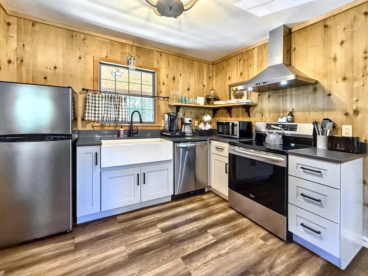 A charming kitchen with a farmhouse sink, stainless steel appliances, wooden paneling, and a window adorned with checkered curtains. The space exudes warmth and functionality, ideal for preparing home-cooked meals or sharing stories over breakfast.