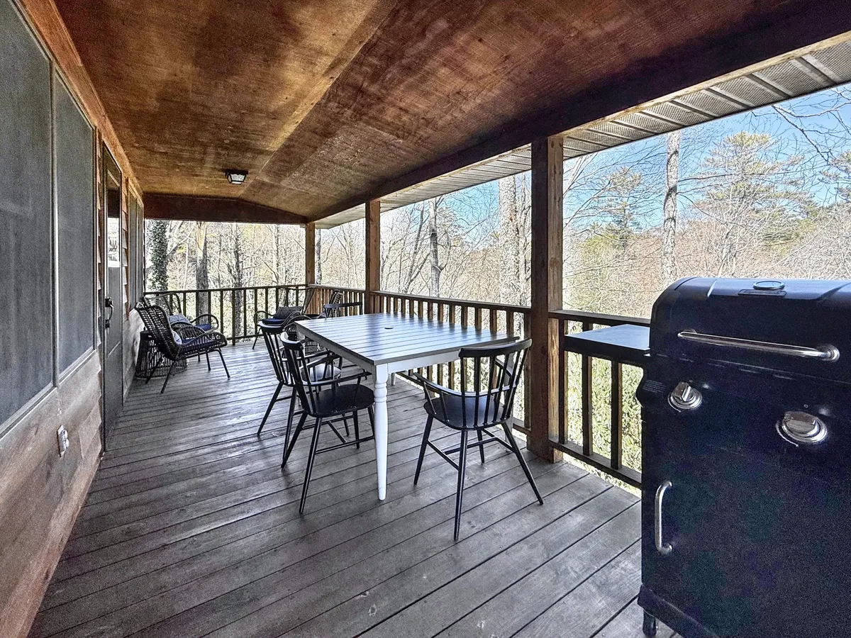 This image highlights a wooden outdoor dining table, black chairs, and a propane grill situated on a deck with a railing, all set against a lush natural backdrop. The scene conveys a comfortable, inviting space for outdoor meals and gatherings, with direct access to nature.