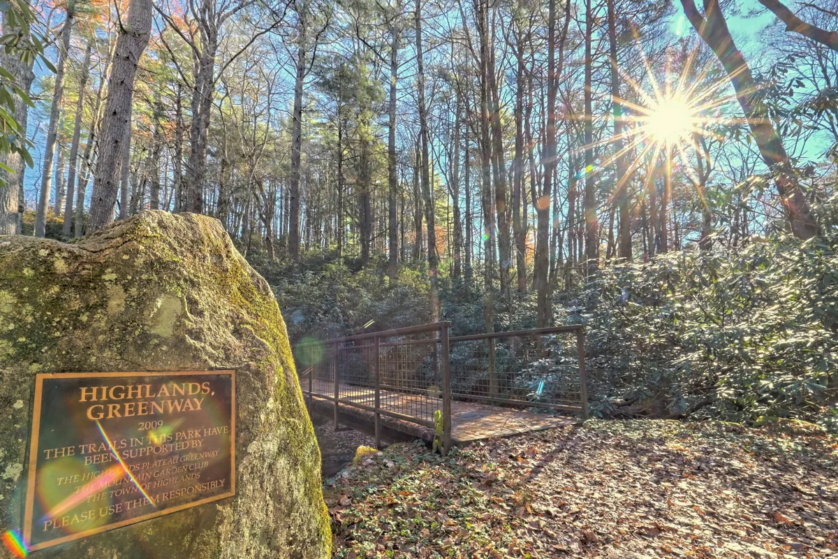 Shows a scenic exterior with a stone sign, a bridge, and sunlight filtering through the trees. The inviting path hints at leisurely walks, connections to nature, and moments to capture the beauty of the Highlands area.