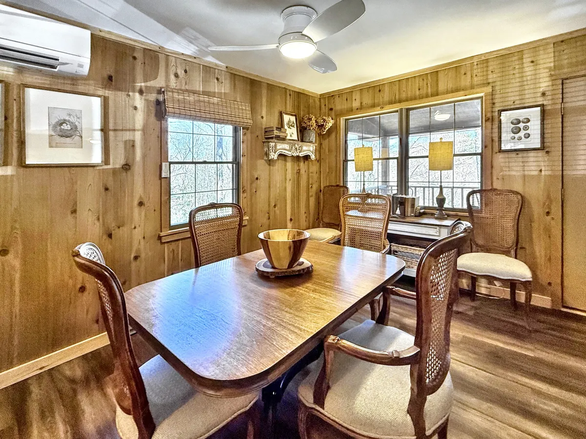 A sunlit dining area with a wooden table, woven chairs, and large windows looking out to the outdoors, suggesting a connection to nature and the changing seasons. The setup encourages family meals, celebrations, and gatherings that shift with the seasons, from spring brunches to summer evening dinners.