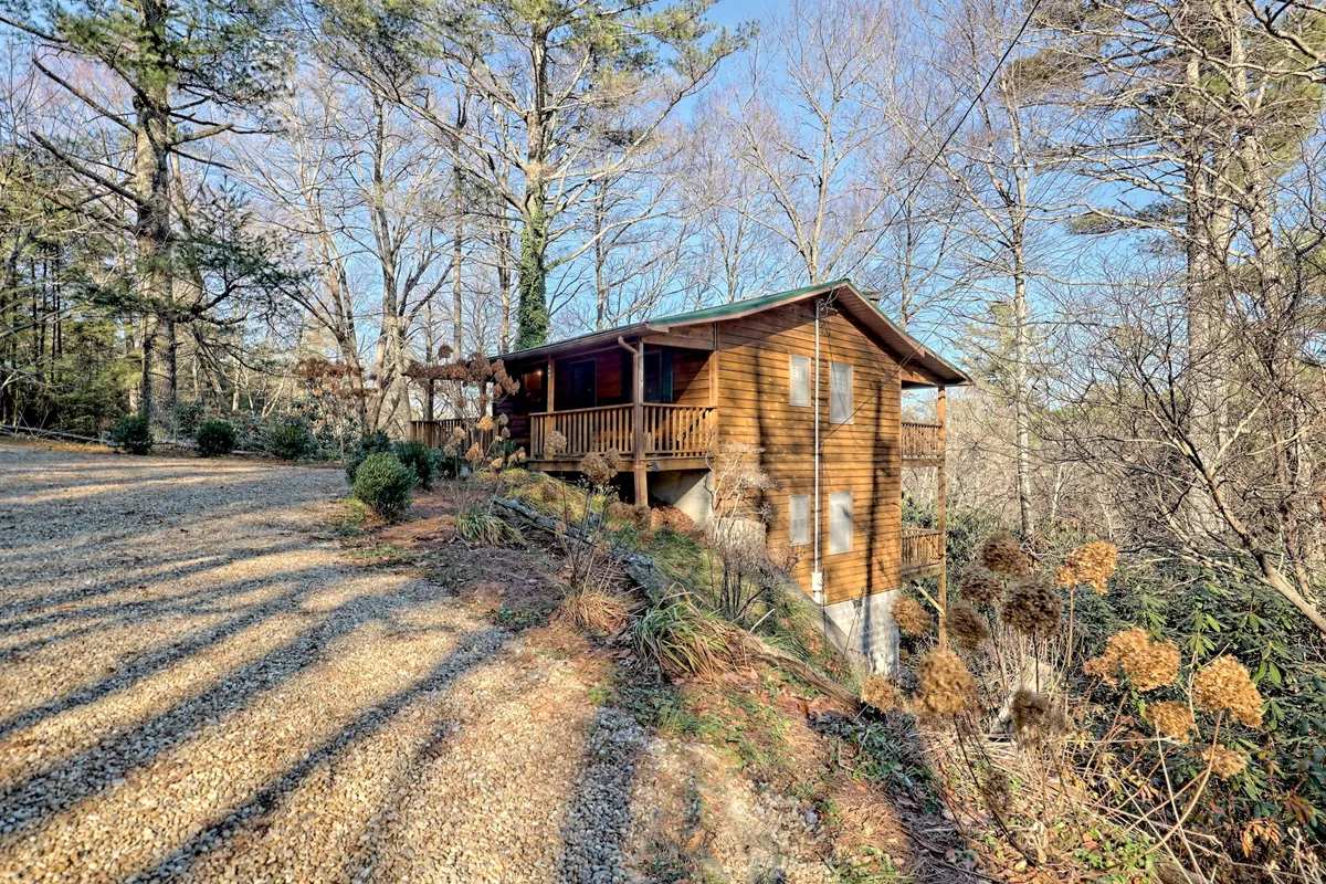 Depicts the charming wooden cabin nestled among surrounding trees, with a gravel driveway leading up to it. The scene suggests a peaceful, natural retreat while maintaining proximity to the action downtown.