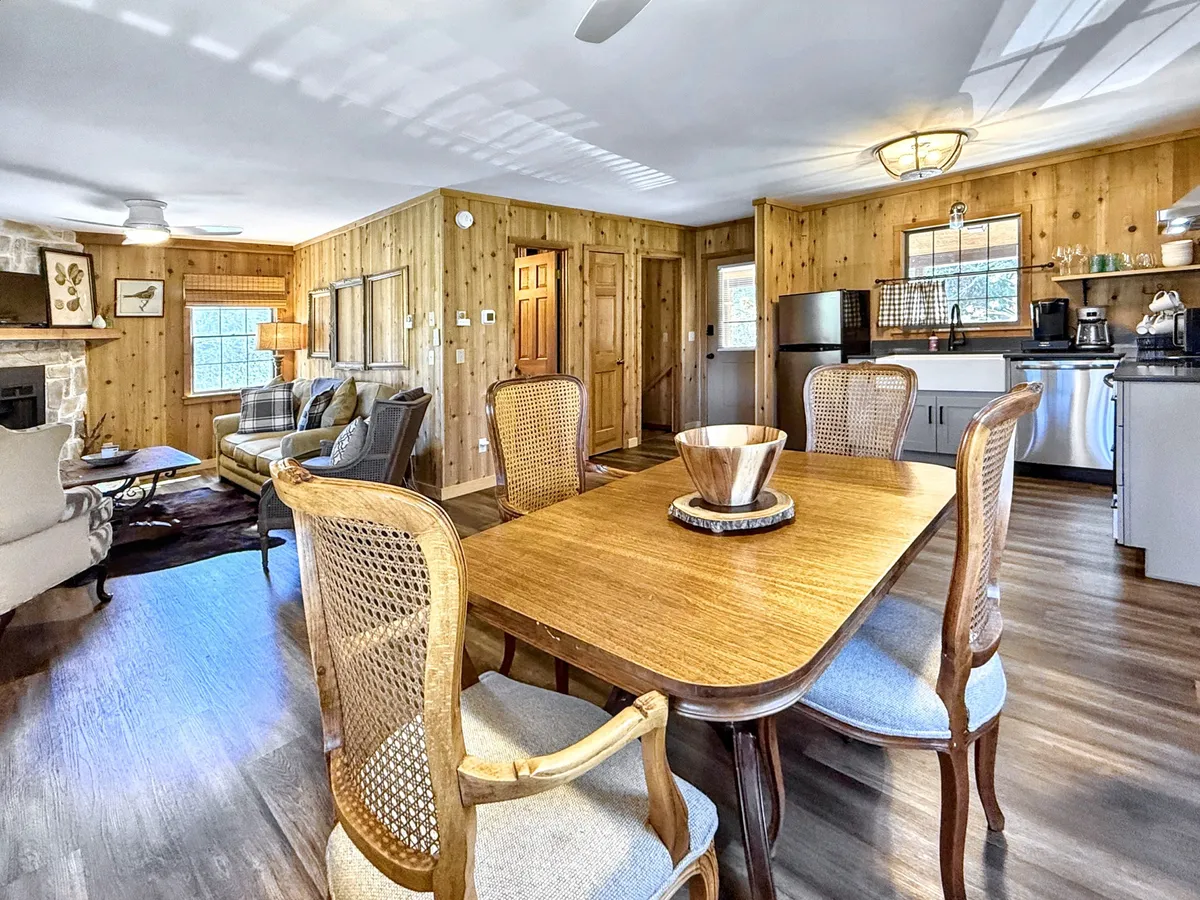 A bright, open indoor space with a wooden dining table, prominent fireplace, and adjacent kitchen appliances. The area is bathed in natural light and accented with rustic wood, creating a harmonious flow between cooking, dining, and lounging spaces.